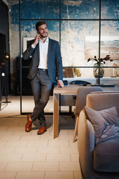 Homem de negócios bonito em terno de pé e falando no smartphone no escritório moderno . — Fotografia de Stock