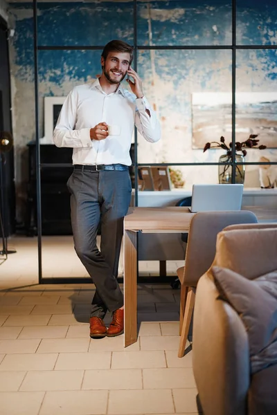 Bel homme d'affaires en costume debout et parlant sur smartphone dans un bureau moderne. — Photo