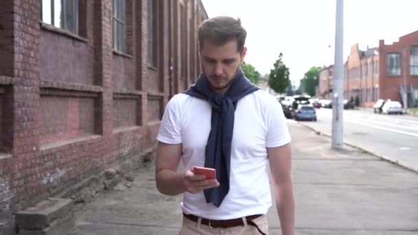 笑顔の男は屋外の携帯電話でメッセージを読む。ハッピーヒップスター男探してスマートフォン画面で都市部の背景 — ストック動画