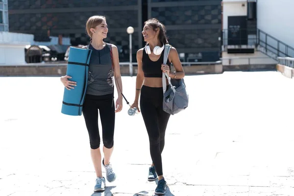 Deux jeunes femmes athlétiques souriantes et heureuses marchant à l'extérieur ensemble après l'entraînement sportif, se parlent et se serrent dans leurs bras. — Photo