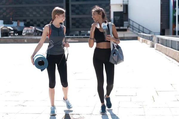 Zwei fröhlich lächelnde junge Sportlerinnen, die nach dem Sport gemeinsam im Freien spazieren gehen, miteinander reden und einander umarmen. — Stockfoto