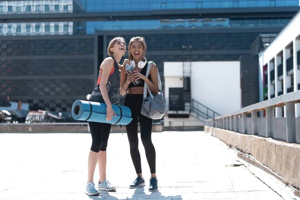 Deux jeunes femmes athlétiques souriantes et heureuses marchant à l'extérieur ensemble après l'entraînement sportif, se parlent et se serrent dans leurs bras. — Photo