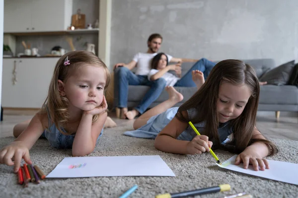 Kinderen zussen spelen samen tekenen op de vloer terwijl jonge ouders ontspannen thuis op de bank, kleine meisjes hebben plezier — Stockfoto