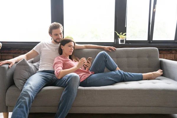 Pareja feliz disfrutando de contenido multimedia en un teléfono inteligente sentado en un sofá en casa. — Foto de Stock