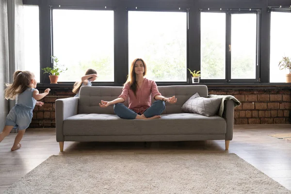 Mujer tranquila concentrándose en ejercicios de yoga en el sofá en casa mientras dos niños ruidosos ríen, corren — Foto de Stock