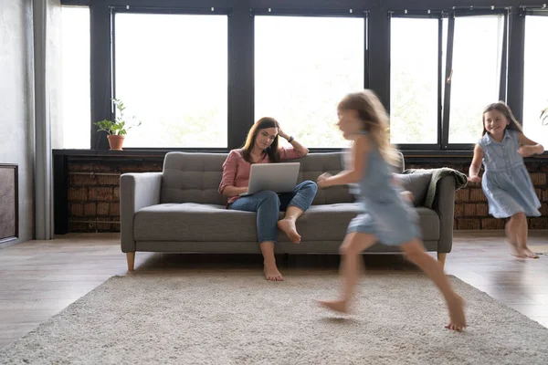 Jeune mère célibataire travaillant sur un ordinateur portable dans un grenier assis sur un canapé tandis que ses filles courent autour d'elle et crient. — Photo