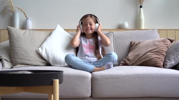Linda niña escuchando música en los auriculares mientras está sentada en el sofá — Vídeo de stock