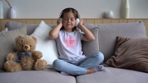 Cute little girl listening to music in headphones while sitting on the couch — Stock Video