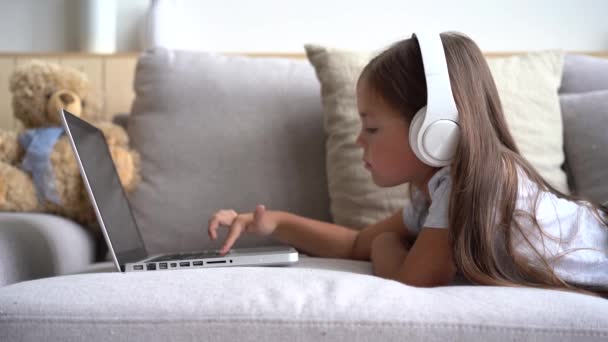 Усміхнена школярка в навушниках за допомогою ноутбука, що лежить на дивані — стокове відео