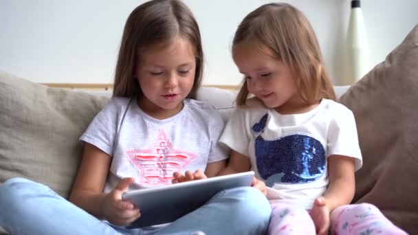 Küçük şirin kız kardeşler yan yana oturup tabletten bir şeyler izliyorlar. — Stok video