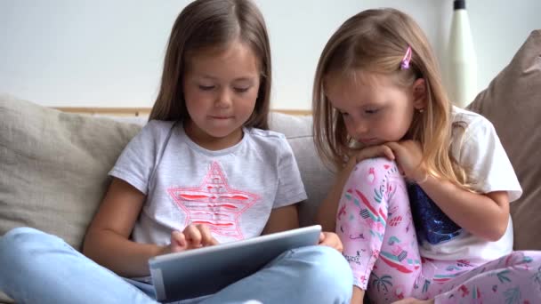 Симпатичні маленькі сестри, сидячи поруч один з одним, дивиться щось на планшет — стокове відео