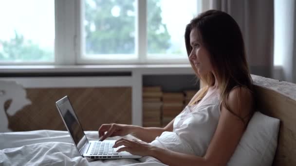 Junge Frau tippt am Laptop und sitzt morgens im Bett — Stockvideo