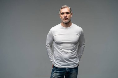 Gri arka planda, kot pantolon ve beyaz tişört giyen orta yaşlı neşeli bir adam..