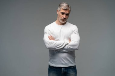 Gri arka planda, kot pantolon ve beyaz tişört giyen orta yaşlı neşeli bir adam..