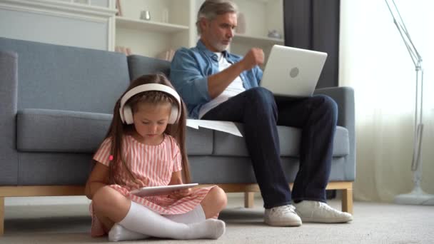 Pai trabalhando com laptop e filha jogando no tablet na sala de estar — Vídeo de Stock