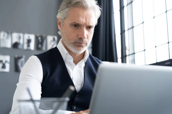 Concentrado en el trabajo. Hombre maduro en traje completo usando el ordenador portátil mientras trabaja en la oficina moderna. —  Fotos de Stock