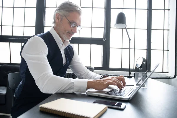 Geconcentreerd op het werk. Volwassen man in vol pak met behulp van laptop tijdens het werken in modern kantoor. — Stockfoto