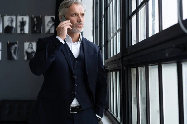 Concepto de éxito. Elegante hombre barbudo maduro en traje elegante hablando en el teléfono inteligente, de pie cerca de la ventana. —  Fotos de Stock