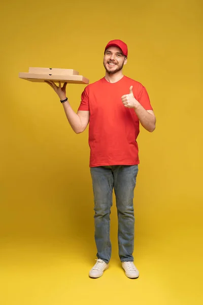 Glücklicher Mann vom Lieferdienst in rotem T-Shirt und Mütze, der Essen bestellt und Pizzakartons isoliert über gelb hält — Stockfoto