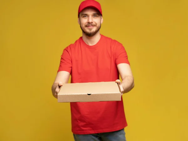 Hombre feliz del servicio de entrega en camiseta roja y gorra dando orden de comida y sosteniendo la caja de pizza aislada sobre amarillo — Foto de Stock