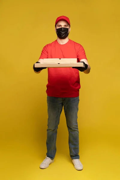 Entrega homem em boné vermelho, t-shirt uniforme, máscara protetora e luvas isoladas em amarelo — Fotografia de Stock