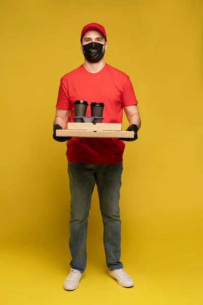 Entrega homem em boné vermelho, t-shirt uniforme, máscara protetora e luvas isoladas em amarelo — Fotografia de Stock