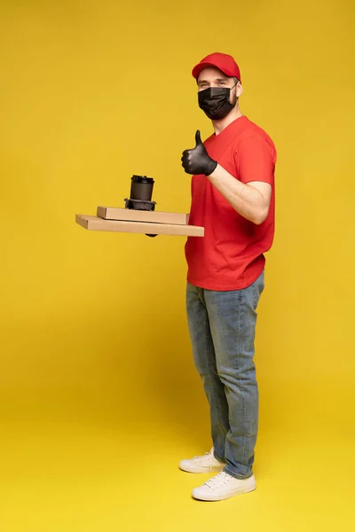 Entrega homem em boné vermelho, t-shirt uniforme, máscara protetora e luvas isoladas em amarelo — Fotografia de Stock