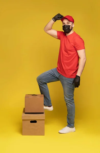 Koruyucu maskeli bir teslimatçı ve sarı arka planda kapalı bir kutuyla eldivenli.. — Stok fotoğraf