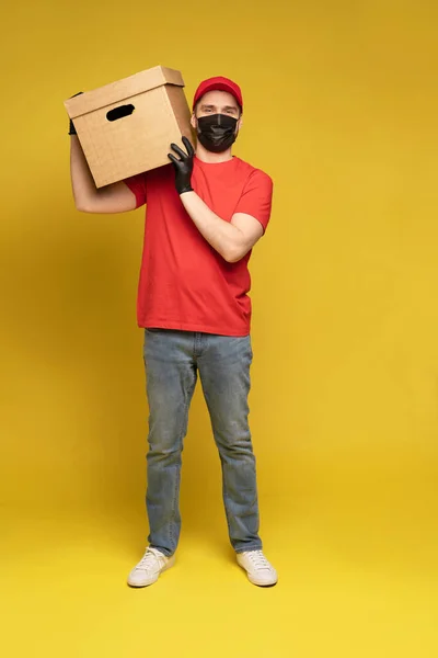 Delivery man in protective mask and gloves with box in studio isolated on yellow background. — Stock Photo, Image