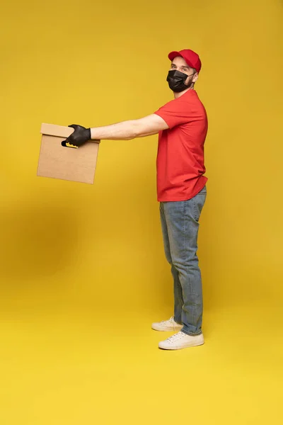 Entrega hombre en máscara protectora y guantes con caja en estudio aislado sobre fondo amarillo. — Foto de Stock