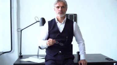 Ofiste duran gri sakallı, ceketli, beyaz bir adam.