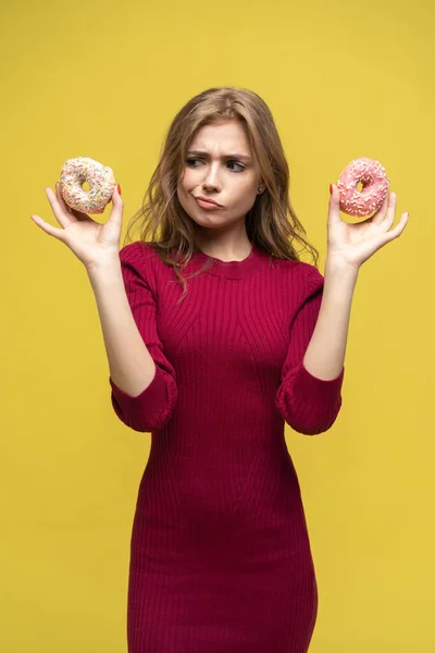Donutlar arasında seçim yapan kafası karışmış genç bir kadın. Sarı arkaplan üzerinde stüdyo resmi. — Stok fotoğraf
