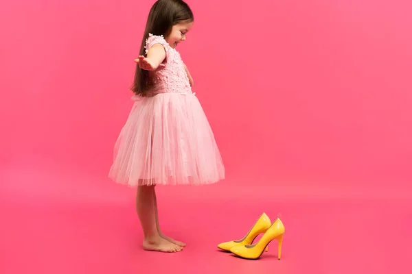 Engraçado criança menina fashionista no vestido vai colocar em grandes mães sapatos amarelos no fundo colorido. — Fotografia de Stock