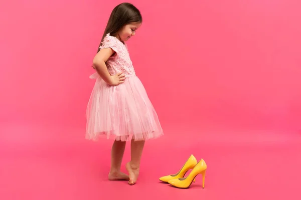 Funny child girl fashionista in dress going to put on big mothers yellow shoes on colored background. — Stock Photo, Image