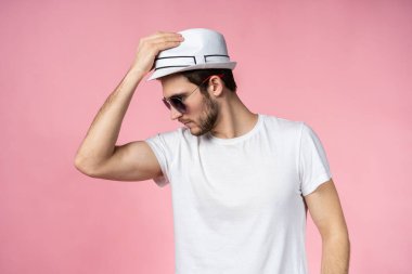 Şapkalı ve güneş gözlüklü neşeli genç adam pembe arka planda duruyor ve gülümsüyor..