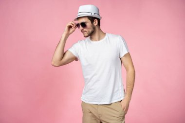 Şapkalı ve güneş gözlüklü neşeli genç adam pembe arka planda duruyor ve gülümsüyor..
