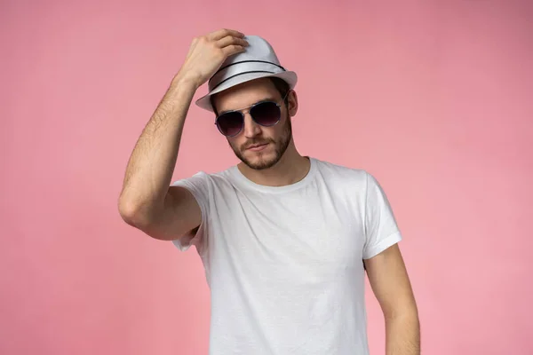 Vidám fiatalember kalapban és napszemüvegben áll és mosolyog rózsaszín háttér felett. — Stock Fotó