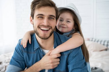 Yakışıklı baba ve tatlı kızının portresi. Sarılmak, kameraya bakmak ve evde kanepede otururken gülümsemek..