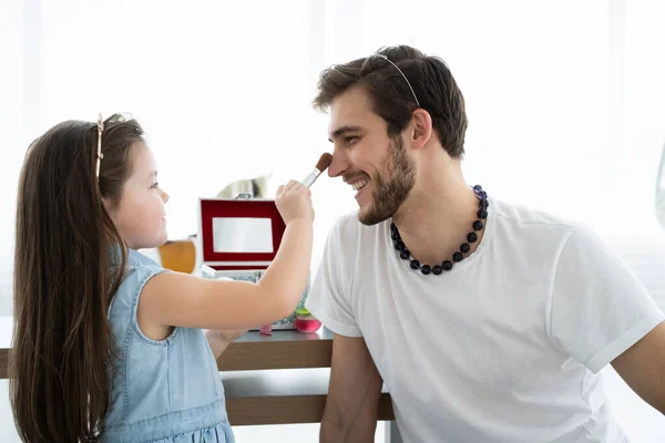 Mignonne petite fille et son beau jeune père en couronnes jouent ensemble dans la chambre des enfants. Fille fait son père un maquillage. — Photo