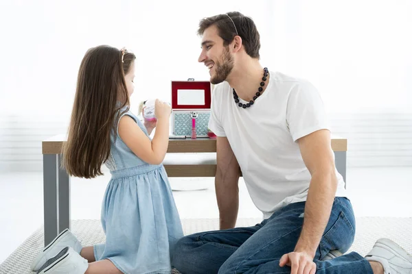 Mignonne petite fille et son beau jeune père en couronnes jouent ensemble dans la chambre des enfants. Fille fait son père un maquillage. — Photo