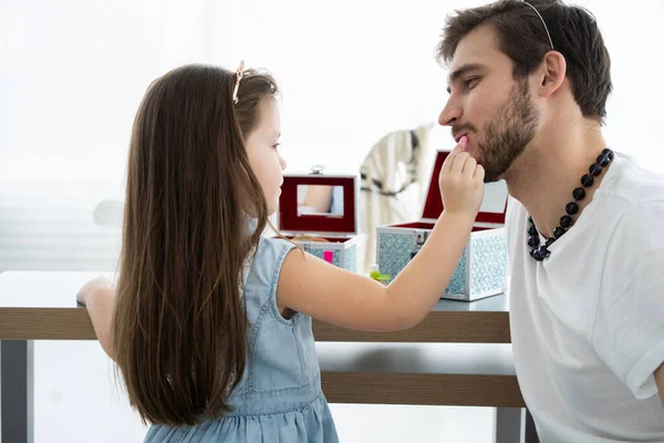 Cute little daughter and her handsome young dad in crowns are playing together in childs room. Girl is doing her dad a makeup. — Stock Photo, Image