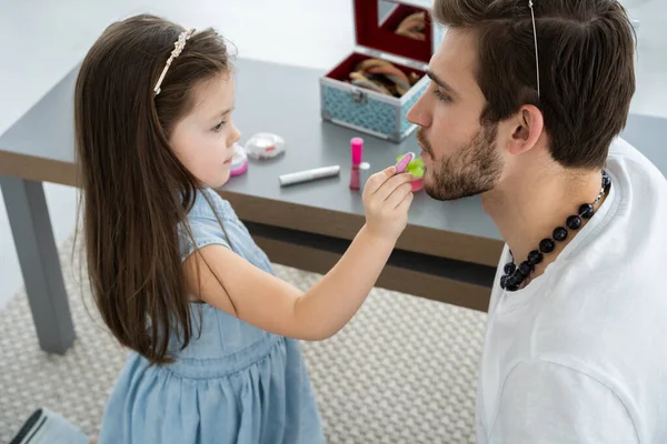 Mignonne petite fille et son beau jeune père en couronnes jouent ensemble dans la chambre des enfants. Fille fait son père un maquillage. — Photo