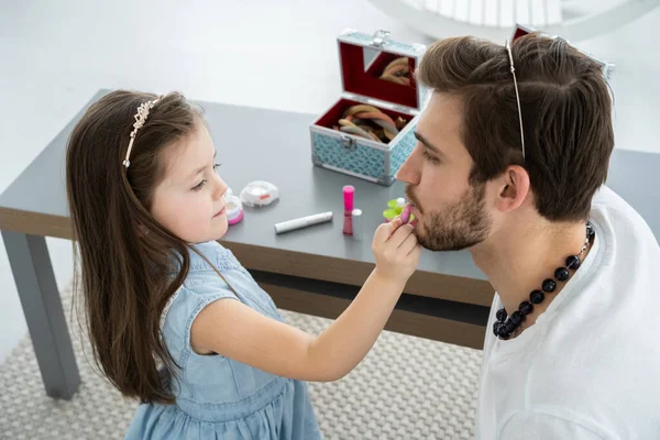 Mignonne petite fille et son beau jeune père en couronnes jouent ensemble dans la chambre des enfants. Fille fait son père un maquillage. — Photo