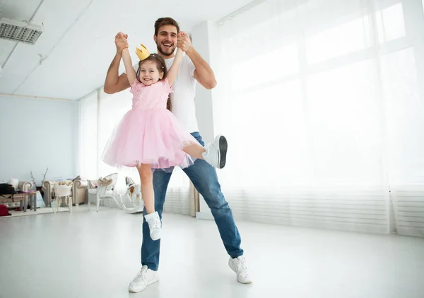 Die süße kleine Tochter und ihr hübscher junger Papa in Kronen spielen zusammen im Kinderzimmer. Mädchen tut ihrem Vater ein Make-up. — Stockfoto