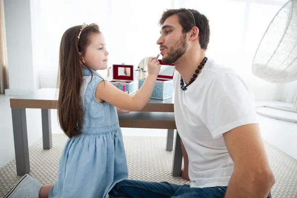 Die süße kleine Tochter und ihr hübscher junger Papa in Kronen spielen zusammen im Kinderzimmer. Mädchen tut ihrem Vater ein Make-up. — Stockfoto