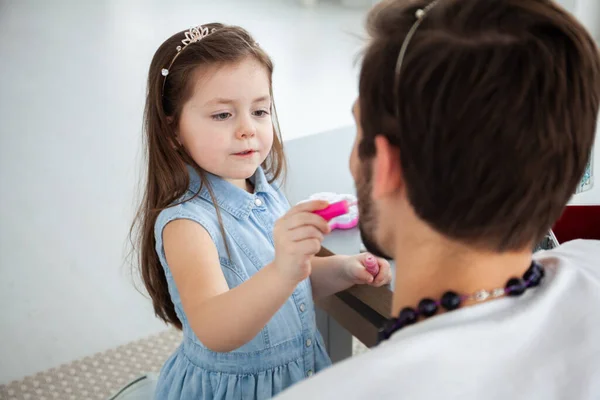 Cute little daughter and her handsome young dad in crowns are playing together in childs room. Girl is doing her dad a makeup. — Stock Photo, Image