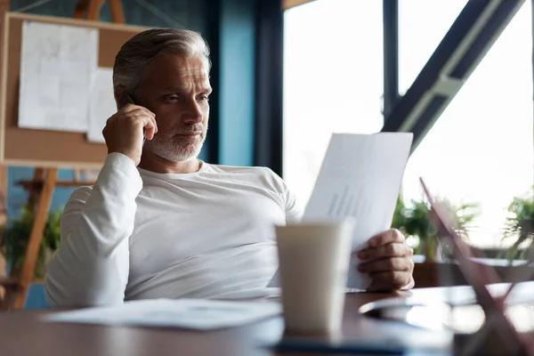 회색 머리의 성숙 한 사업가가 책상에 앉아 컴퓨터를 하고 스마트폰으로 이야기하고 있습니다. 전화 통화. — 스톡 사진