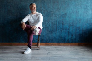 Koyu mavi arka planda, sandalyede oturan gri saçlı, olgun, yakışıklı bir adamın portresi.