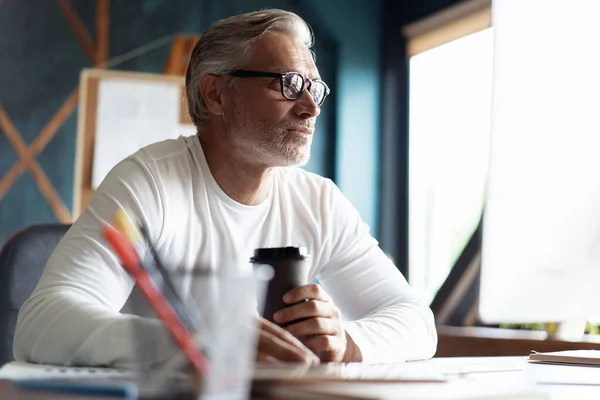 Casual de cabelos grisalhos homem maduro com café trabalhando na mesa no escritório criativo ocupado. — Fotografia de Stock
