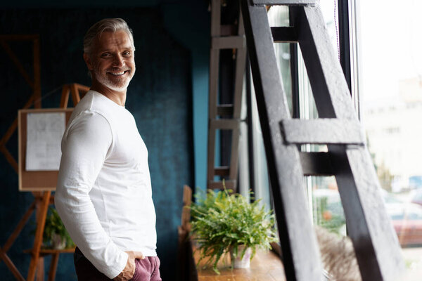 Smiling attractive Casual Grey-haired mature man standing near window looking at the camera.